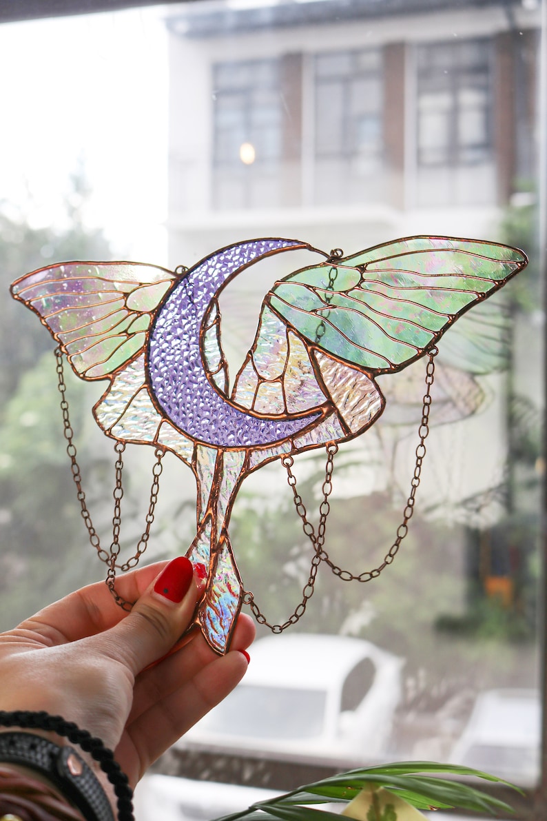 May include: A stained glass window hanging with a crescent moon and iridescent butterfly wings. The wings are green, purple, and white. The moon is purple and the hanging chains are copper.