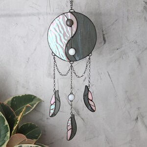 May include: A stained glass yin yang symbol with pink and gray stripes hangs from a chain with three white beads and three pink and gray feathers.