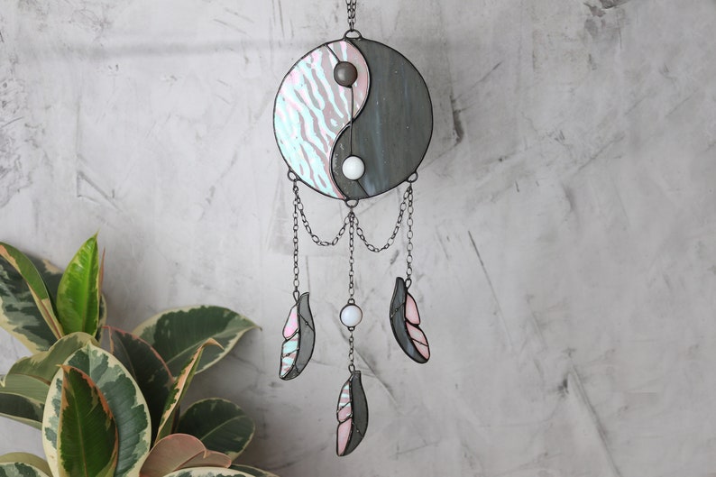 May include: A stained glass yin yang symbol with pink and gray stripes hangs from a chain with three white beads and three pink and gray feathers.