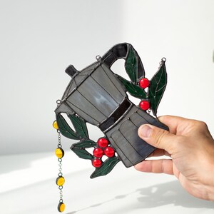 Puede incluir: Un atrapasoles de vidrieras con forma de cafetera moka, con paneles de vidrio gris, hojas de color verde oscuro y bayas de color rojo brillante. Una peque&ntilde;a cadena plateada con cuatro cuentas de vidrio circulares amarillas cuelga del lado.