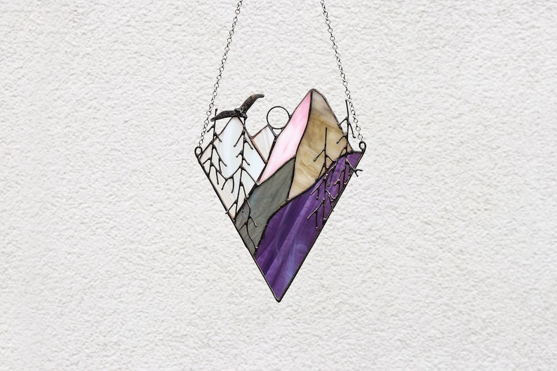 May include: A stained glass window hanging with a chain. The window depicts a mountain range with a pink, brown, gray, and purple color scheme. A bird is flying over the mountains.