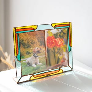Può includere: Una cornice portafoto doppia in vetro colorato con un design colorato. La cornice presenta un bordo azzurro, giallo, arancione e verde acqua. Un lato contiene una foto di un cane e l'altro lato tulipani rossi.