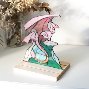 Op de afbeelding: Gebrandschilderde glazen drakenbeeld in roze, groen en blauw, gemonteerd op een houten voet. De draak heeft uitgespreide vleugels en zit op een kleurrijke, gestileerde berg. Een decoratief item.