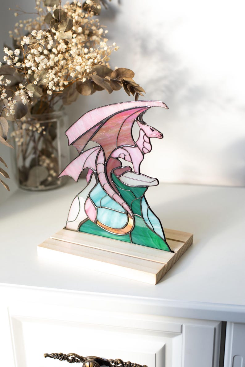 Op de afbeelding: Gebrandschilderde glazen drakenbeeld in roze, groen en blauw, gemonteerd op een houten voet. De draak heeft uitgespreide vleugels en zit op een kleurrijke, gestileerde berg. Een decoratief item.