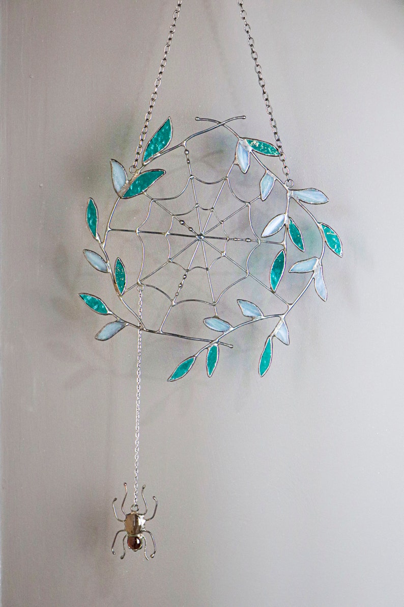 May include: A silver metal spider web with teal and white glass leaves hanging from a silver chain. A silver spider charm hangs from the bottom of the web.