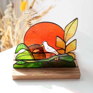 Op de afbeelding: Gebrandschilderd glaskunstwerk met een groene alligator met een oranje zonsondergang, een witte vogel en gele en groene bladeren. Het stuk is gemonteerd op een houten voet, een uniek decoratief item.