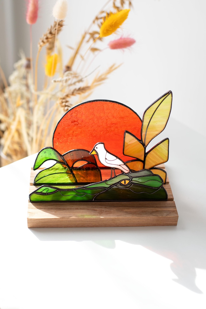 Op de afbeelding: Gebrandschilderd glaskunstwerk met een groene alligator met een oranje zonsondergang, een witte vogel en gele en groene bladeren. Het stuk is gemonteerd op een houten voet, een uniek decoratief item.