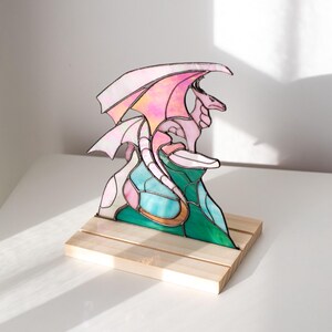 Op de afbeelding: Een gebrandschilderde drakenbeeldhouwwerk met roze en witte vleugels, een roze lichaam en een turquoise en groene basis. De draak is gemonteerd op een kleine, natuurlijke houten basis. De sculptuur wordt op een wit oppervlak getoond.
