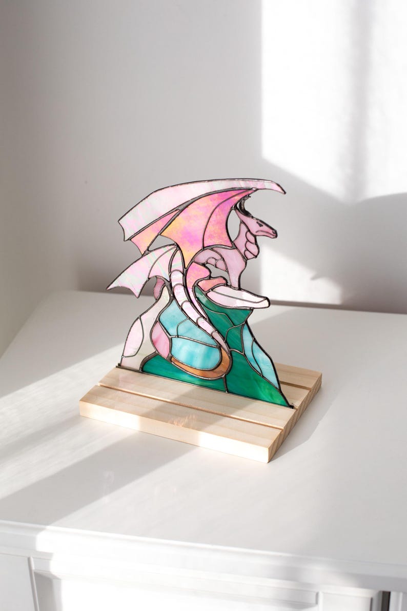 Op de afbeelding: Een gebrandschilderde drakenbeeldhouwwerk met roze en witte vleugels, een roze lichaam en een turquoise en groene basis. De draak is gemonteerd op een kleine, natuurlijke houten basis. De sculptuur wordt op een wit oppervlak getoond.