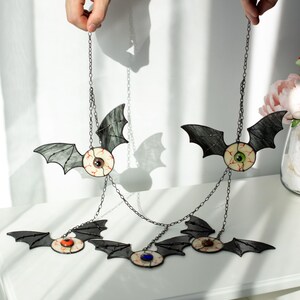 May include: A hanging mobile featuring five stained glass bat figures, each with a central eyeball design. The bats have dark grey wings and irises in various colours including green, blue, orange, and brown, all connected by thin metal chains.