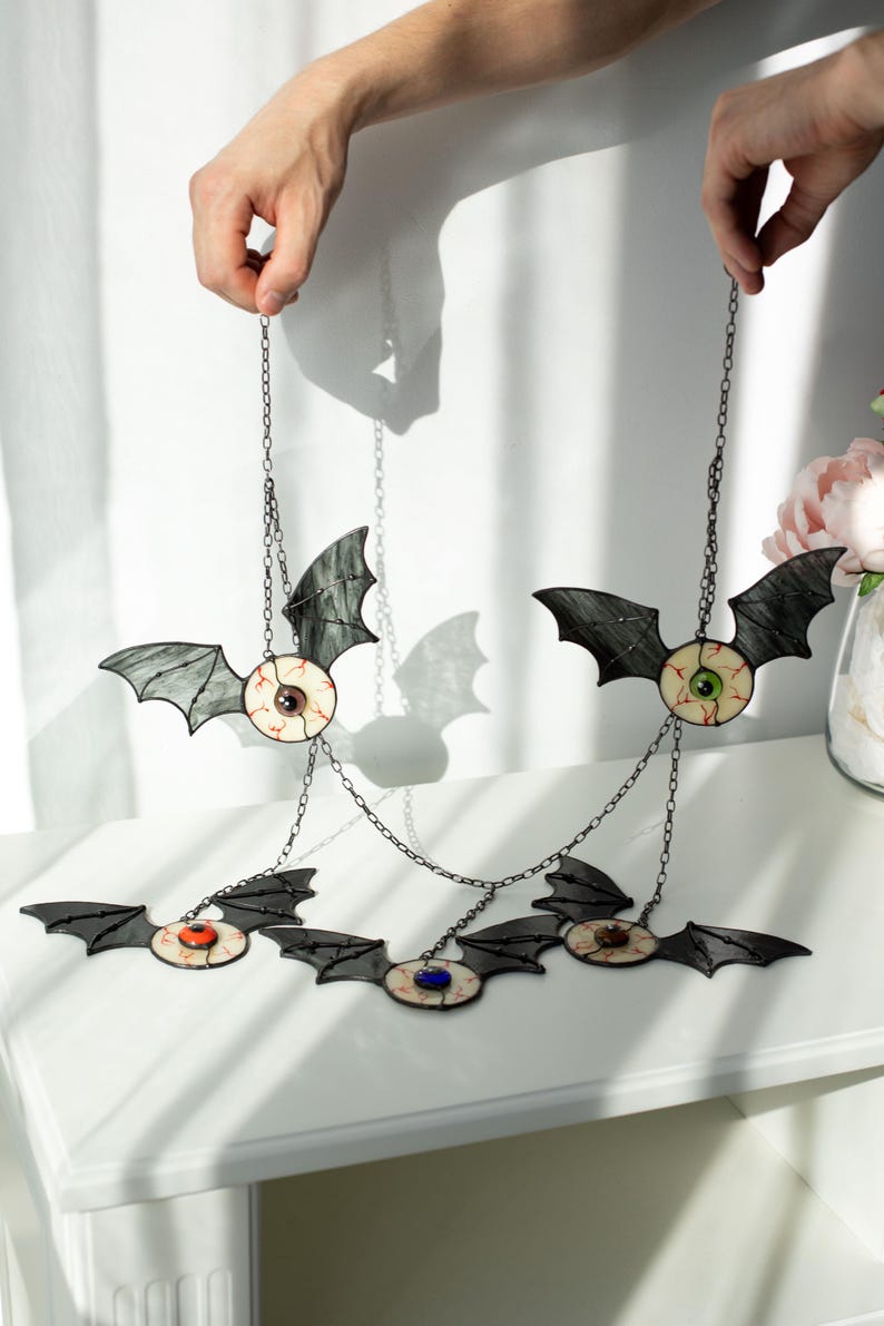 May include: A hanging mobile featuring five stained glass bat figures, each with a central eyeball design. The bats have dark grey wings and irises in various colours including green, blue, orange, and brown, all connected by thin metal chains.