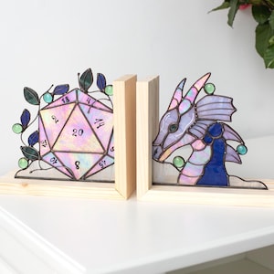 May include: Stained glass bookends featuring a 20-sided die and a dragon head. The die is iridescent with a pink and purple hue, while the dragon is shades of purple and blue. Both are set on wooden bases.