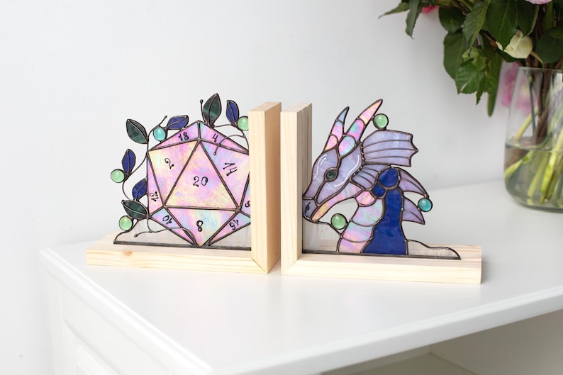 May include: Stained glass bookends featuring a 20-sided die and a dragon head. The die is iridescent with a pink and purple hue, while the dragon is shades of purple and blue. Both are set on wooden bases.
