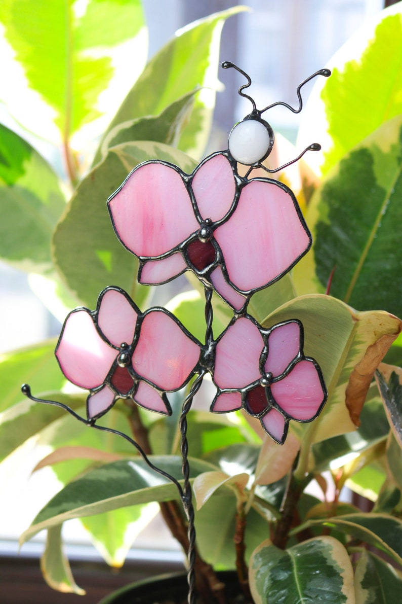 K&ouml;nnte beinhalten: Eine rosafarbene Orchideenbl&uuml;te aus Glasmalerei mit einer wei&szlig;en Perle an der Spitze des Stiels. Die Orchidee besteht aus Glas und Draht und ist ein Dekorationsst&uuml;ck.