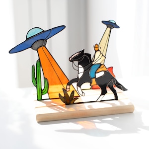 May include: A stained glass sculpture depicting a cowboy on horseback being abducted by a UFO. The scene includes a cactus, desert plants, and two UFOs with orange and yellow beams. The sculpture is mounted on a wooden base.
