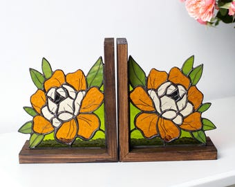Puede incluir: Juego de dos sujetalibros de madera con diseños florales de vidrieras. Cada sujetalibros tiene una base y un marco de madera marrón oscuro, con un diseño floral naranja y blanco y hojas verdes. El fondo es verde brillante.