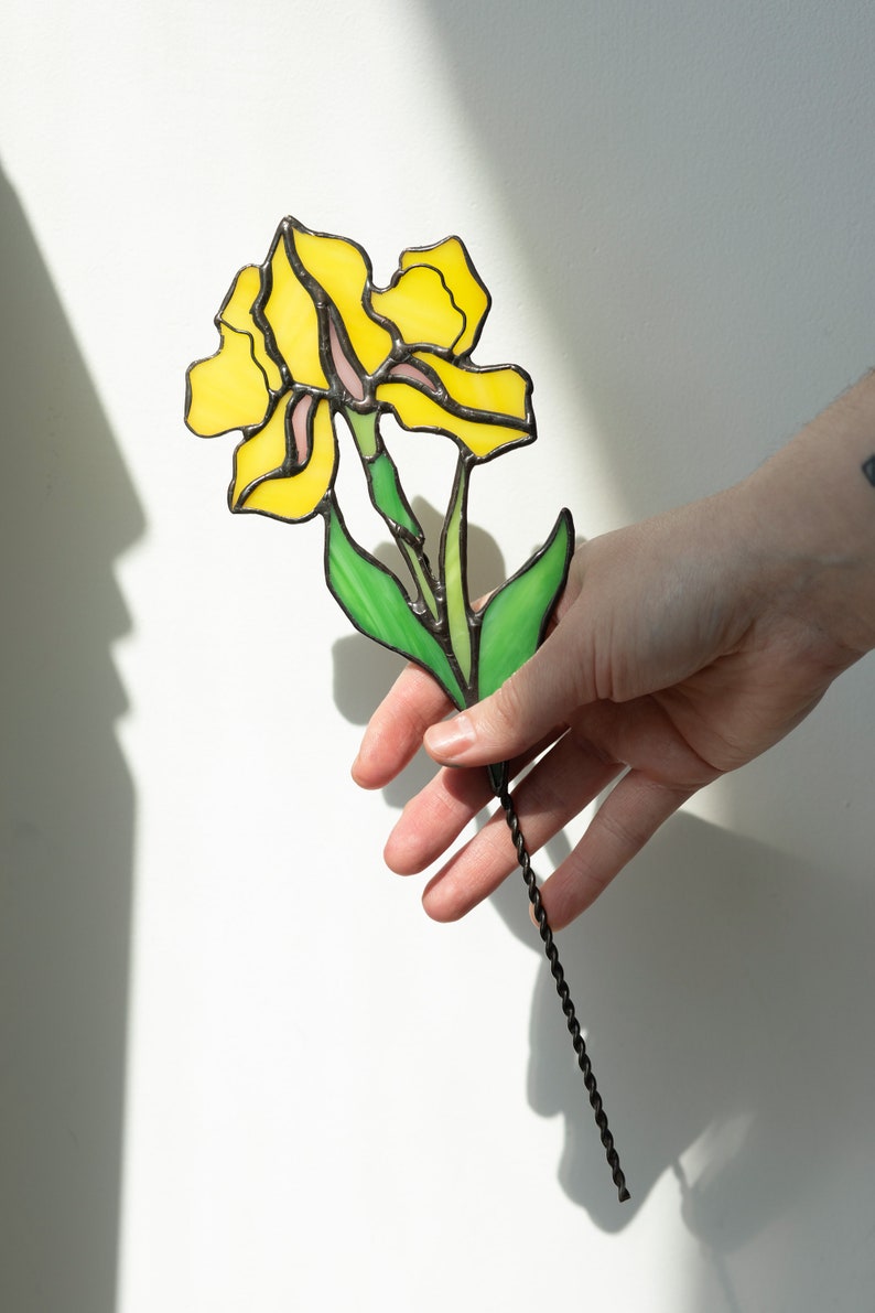 May include: A stained glass flower with yellow petals and green leaves, attached to a black metal stem.