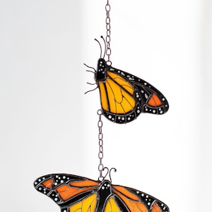 May include: Stained glass monarch butterfly suncatcher with orange and black wings, white spots, and a brown chain. The butterfly is suspended from a green leaf-shaped piece.