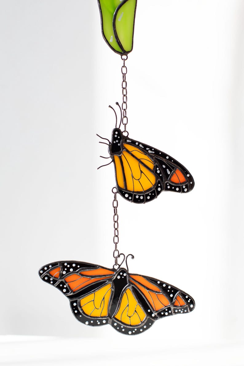 May include: Stained glass monarch butterfly suncatcher with orange and black wings, white spots, and a brown chain. The butterfly is suspended from a green leaf-shaped piece.