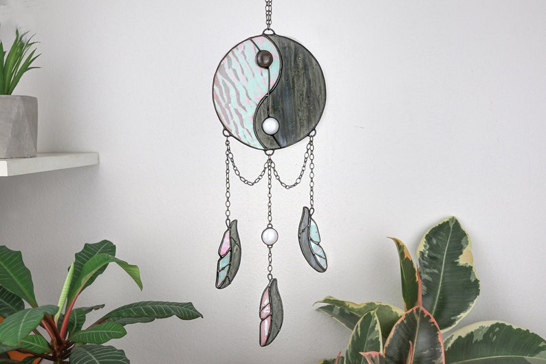 May include: A stained glass yin yang dream catcher with a light blue and gray design. The dream catcher has three feathers hanging from it, two are light blue and gray and one is pink and gray.