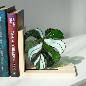 K&ouml;nnte beinhalten: Ein Buchst&uuml;tze in Form eines Monstera-Blattes aus Buntglas in Gr&uuml;n- und Wei&szlig;t&ouml;nen mit einem Holzsockel. Die Buchst&uuml;tze ist zum Halten von B&uuml;chern konzipiert und steht neben einer Reihe von B&uuml;chern auf einem wei&szlig;en Regal.