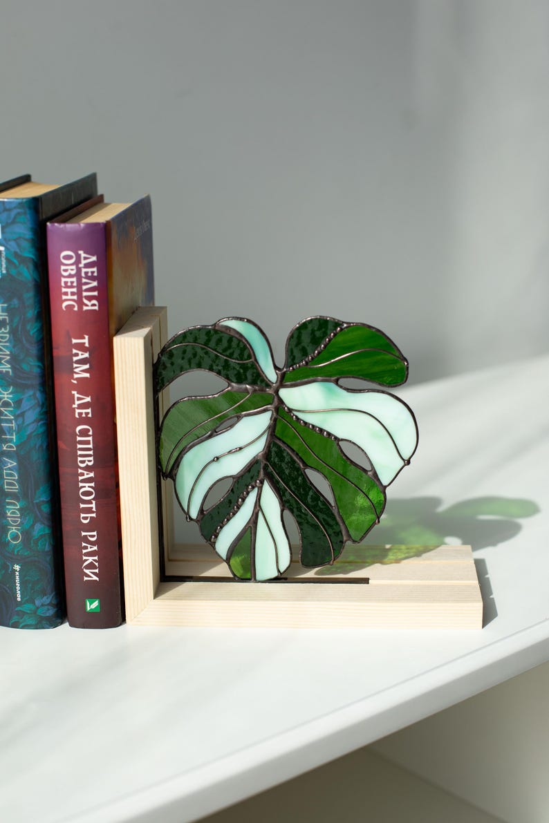 K&ouml;nnte beinhalten: Ein Buchst&uuml;tze in Form eines Monstera-Blattes aus Buntglas in Gr&uuml;n- und Wei&szlig;t&ouml;nen mit einem Holzsockel. Die Buchst&uuml;tze ist zum Halten von B&uuml;chern konzipiert und steht neben einer Reihe von B&uuml;chern auf einem wei&szlig;en Regal.