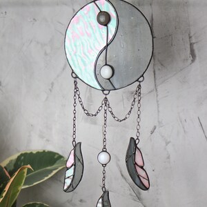 May include: A stained glass yin yang symbol with pink and blue swirls hangs from a chain with three gray and pink feathers and white beads.