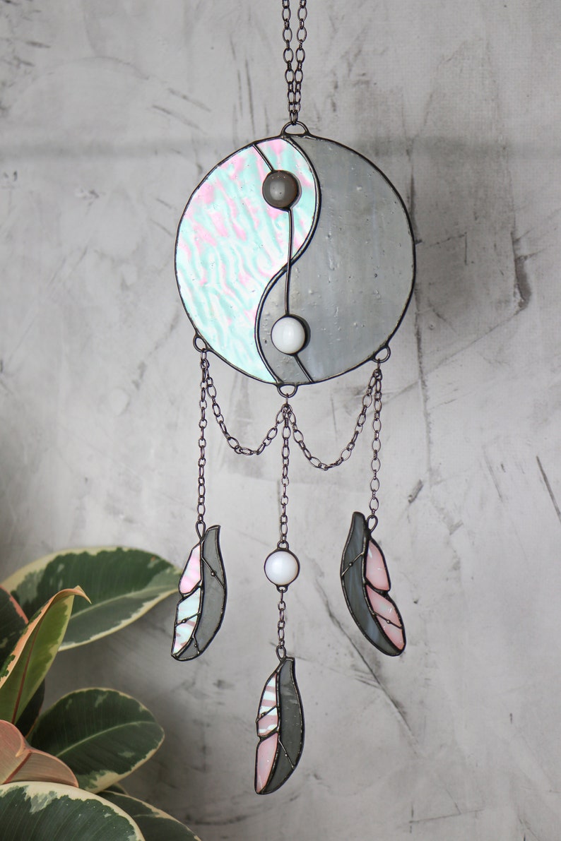 May include: A stained glass yin yang symbol with pink and blue swirls hangs from a chain with three gray and pink feathers and white beads.