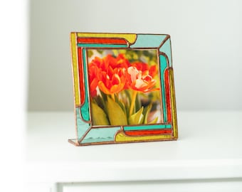 Könnte beinhalten: Ein farbenfroher Glasmalerei-Fotorahmen mit einem Bild von orangefarbenen Tulpen. Der quadratische Rahmen hat einen kupferfarbenen Rand und Abschnitte aus gelbem, türkisem und rotem Glas. Er steht auf einer weißen Oberfläche.