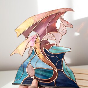 Op de afbeelding: Gebrandschilderde glazen drakenbeeld in tinten roze, paars, blauw en goud. De draak zit op een rotsformatie met een koperen frame. De vleugels zijn gespreid en de draak heeft rode ogen. De basis is zwart.