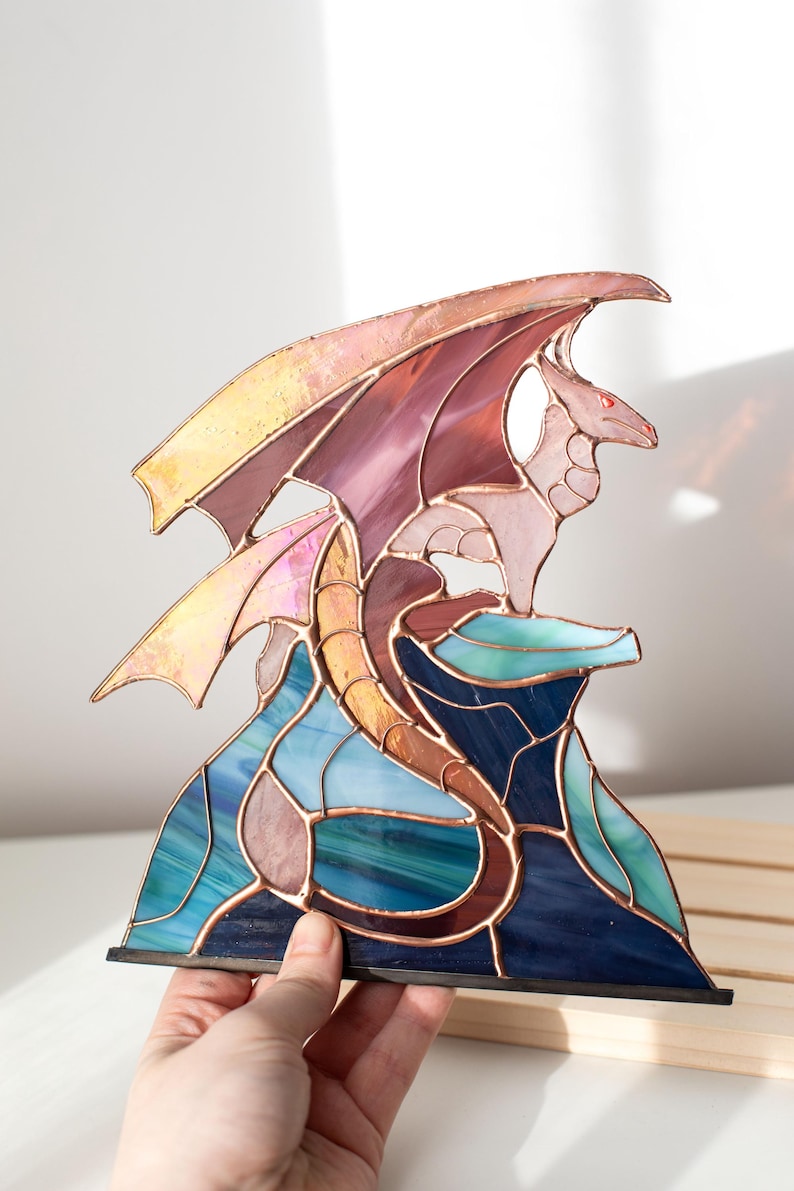 Op de afbeelding: Gebrandschilderde glazen drakenbeeld in tinten roze, paars, blauw en goud. De draak zit op een rotsformatie met een koperen frame. De vleugels zijn gespreid en de draak heeft rode ogen. De basis is zwart.