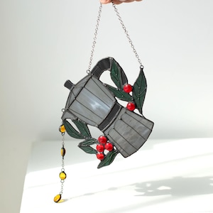 Puede incluir: Un atrapasoles de vidrieras con forma de cafetera moka en tonos grises, decorado con hojas verdes y bayas rojas. Una peque&ntilde;a cadena con cuentas de vidrio amarillas cuelga del pico y una cadena de metal est&aacute; unida para colgar el objeto.