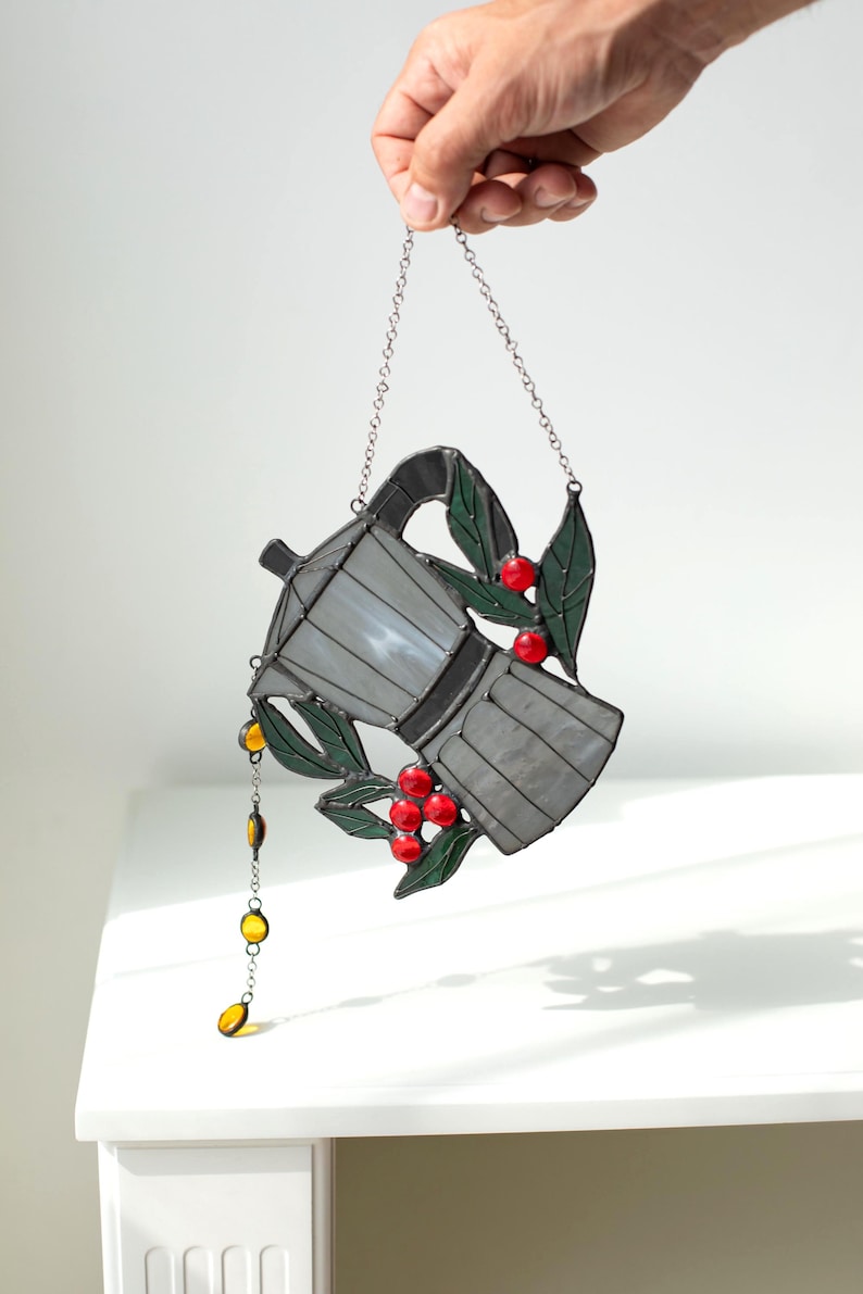 Puede incluir: Un atrapasoles de vidrieras con forma de cafetera moka en tonos grises, decorado con hojas verdes y bayas rojas. Una peque&ntilde;a cadena con cuentas de vidrio amarillas cuelga del pico y una cadena de metal est&aacute; unida para colgar el objeto.