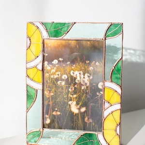 May include: A rectangular stained glass picture frame with a nature scene inside. The frame has a copper border and features yellow, green, and light blue geometric designs. The frame is on a white surface.