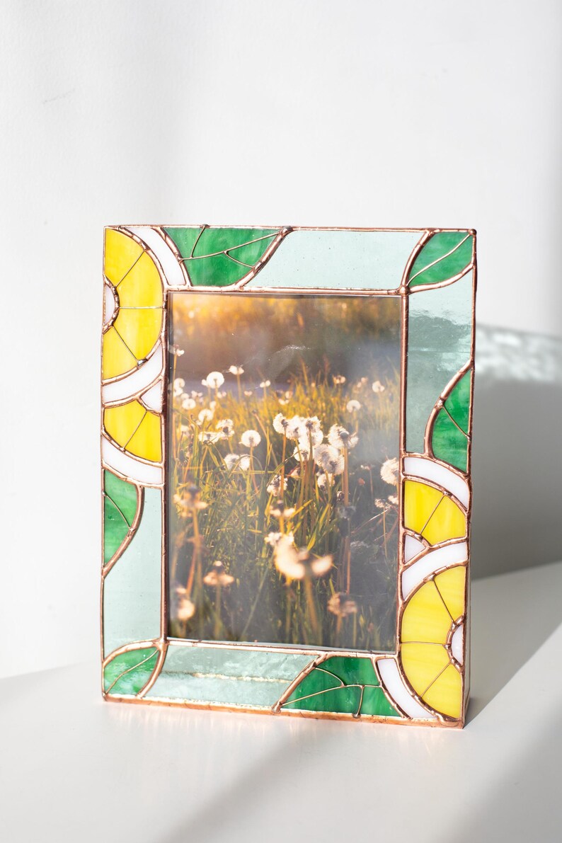 May include: A rectangular stained glass picture frame with a nature scene inside. The frame has a copper border and features yellow, green, and light blue geometric designs. The frame is on a white surface.