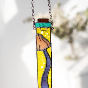 May include: A stained glass pendant shaped like a vial with a yellow background, a blue wavy line, and a brown and orange object inside. The vial is hanging from a silver chain.