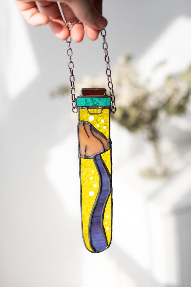 May include: A stained glass pendant shaped like a vial with a yellow background, a blue wavy line, and a brown and orange object inside. The vial is hanging from a silver chain.