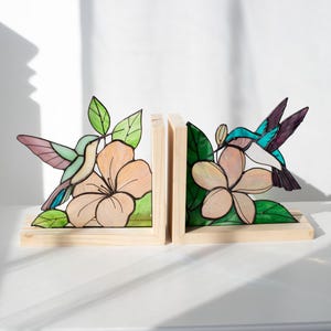 Pu&ograve; includere: Coppia di fermalibri in legno con disegni di colibr&igrave; e fiori in vetro colorato. I fermalibri hanno una base in legno naturale e raffigurano un colibr&igrave; con dettagli rosa, blu e verdi, vicino a un fiore color pesca con foglie verdi.