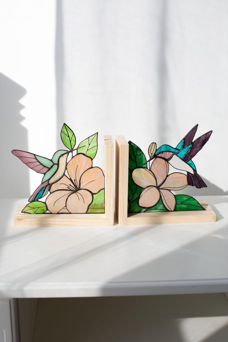 Pu&ograve; includere: Coppia di fermalibri in legno con disegni di colibr&igrave; e fiori in vetro colorato. I fermalibri hanno una base in legno naturale e raffigurano un colibr&igrave; con dettagli rosa, blu e verdi, vicino a un fiore color pesca con foglie verdi.
