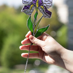 May include: A stained glass iris flower with purple and green glass pieces. The flower is attached to a silver wire stem.