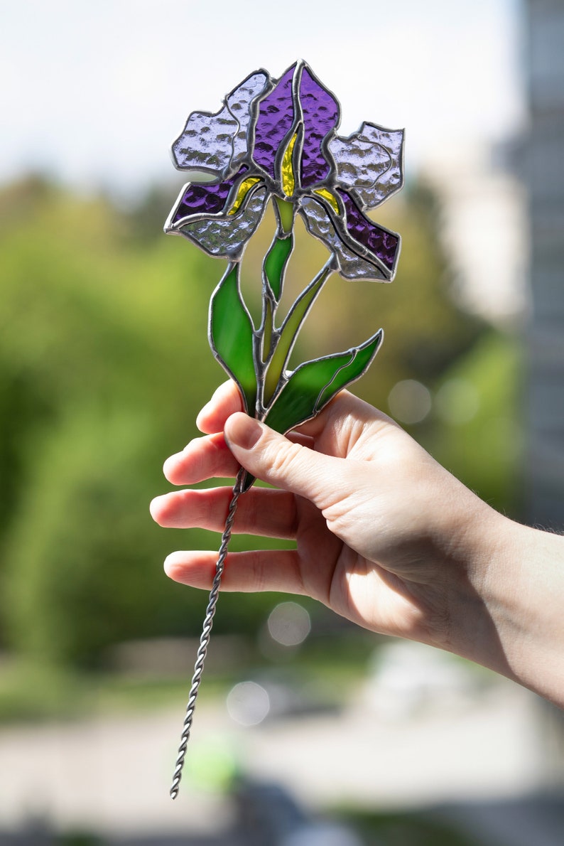 May include: A stained glass iris flower with purple and green glass pieces. The flower is attached to a silver wire stem.
