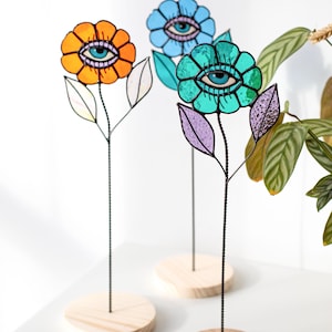 May include: Three colorful stained glass flower sculptures with eye designs. The flowers are orange, blue, and teal, each with a detailed eye in the center. They have black wire stems and sit on round wooden bases, perfect for home decor.