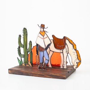 May include: A stained glass cowboy figurine with a white horse, a cactus, and a sunset background. The figurine is mounted on a wooden base.