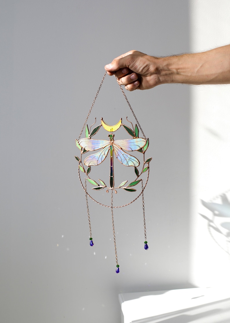 May include: A stained glass dragonfly suncatcher with iridescent wings, green leaves, and a yellow crescent moon. The suncatcher hangs from a copper chain with three blue glass beads.