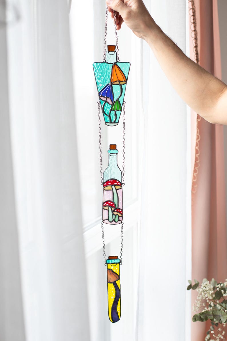May include: A stained glass window hanging with three glass bottles, each containing a different design of mushrooms. The bottles are connected by a silver chain.