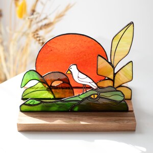 Op de afbeelding: Gebrandschilderd glaskunstwerk met een witte vogel op een groene alligator, tegen een oranje zon. Het stuk bevat gele en groene bladeren en rust op een houten voet. Een decoratief item, ideaal voor weergave.