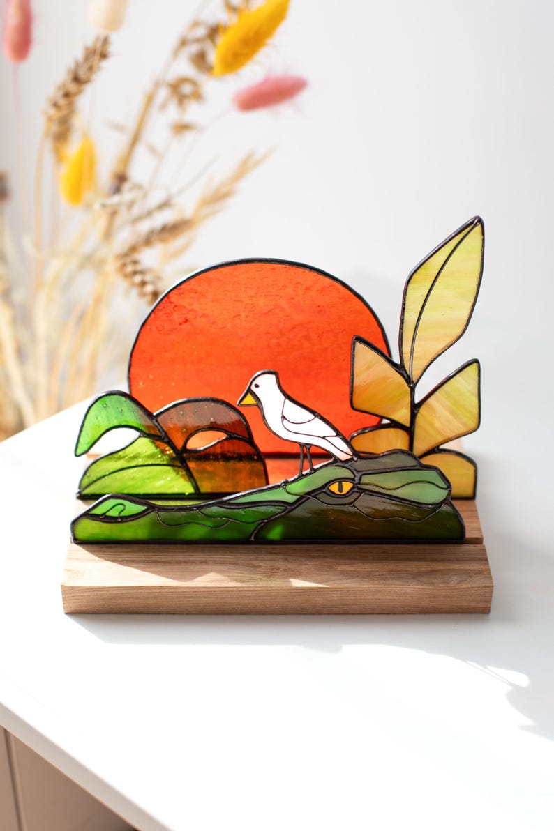 Op de afbeelding: Gebrandschilderd glaskunstwerk met een witte vogel op een groene alligator, tegen een oranje zon. Het stuk bevat gele en groene bladeren en rust op een houten voet. Een decoratief item, ideaal voor weergave.