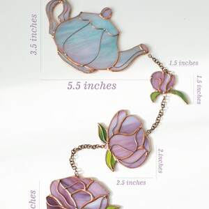 Puede incluir: Un adorno colgante de vitral con una tetera y tres flores conectadas por cadenas de cobre. La tetera mide 8,9 cm por 14 cm. Las flores var&iacute;an de 3,8 cm a 8,9 cm de altura y de 3,8 cm a 7,6 cm de ancho.