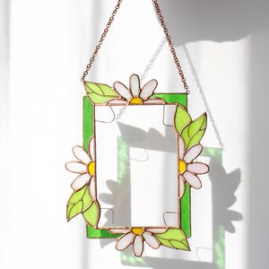 May include: A stained glass frame with a green border, daisy accents, and a copper chain for hanging. The daisies have white petals and yellow centers, with green leaves. The frame is held by a hand.