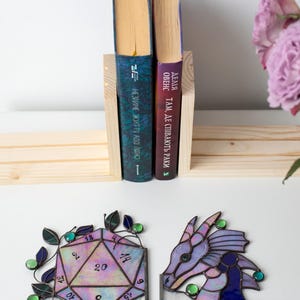 May include: A wooden bookend holding two books with blue and purple covers. Below, two stained glass bookends: a D20 die and a dragon head, both with purple, green, and blue accents. The die has numbers and the dragon has a detailed design.